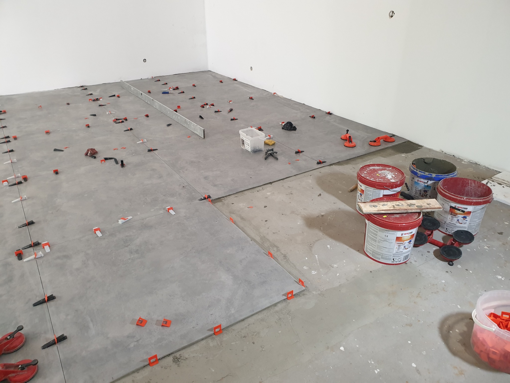 Układanie szarych płytek podłogowych z użyciem systemu poziomowania płytek; widoczne wiadra z klejem Imprefarb, poziomica i narzędzia glazurnicze w surowym wnętrzu.