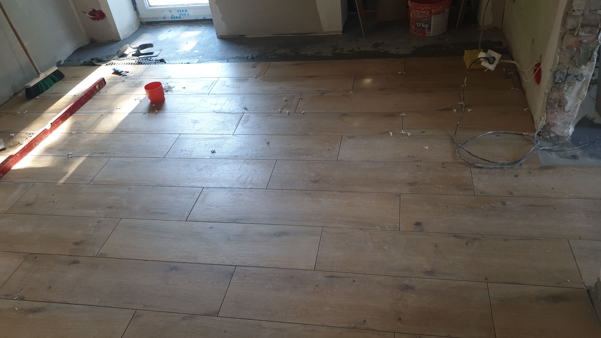 Układanie podłogi z płytek imitujących drewno, widoczne krzyżyki dystansowe, poziomica, wiadro z klejem i fragment ściany w trakcie prac.