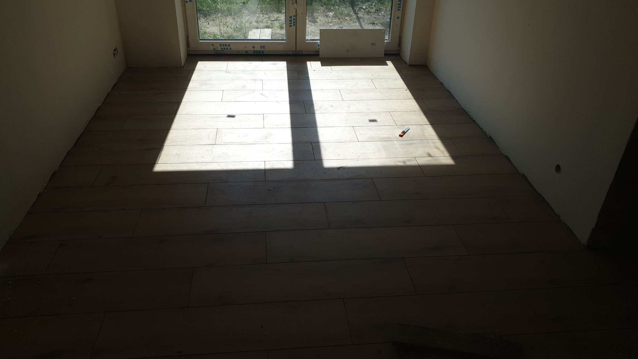 Podłoga wyłożona płytkami imitującymi drewno w nowym pomieszczeniu, z wyraźnym promieniem światła padającym przez okno i narzędziem do cięcia płytek leżącym na podłodze.