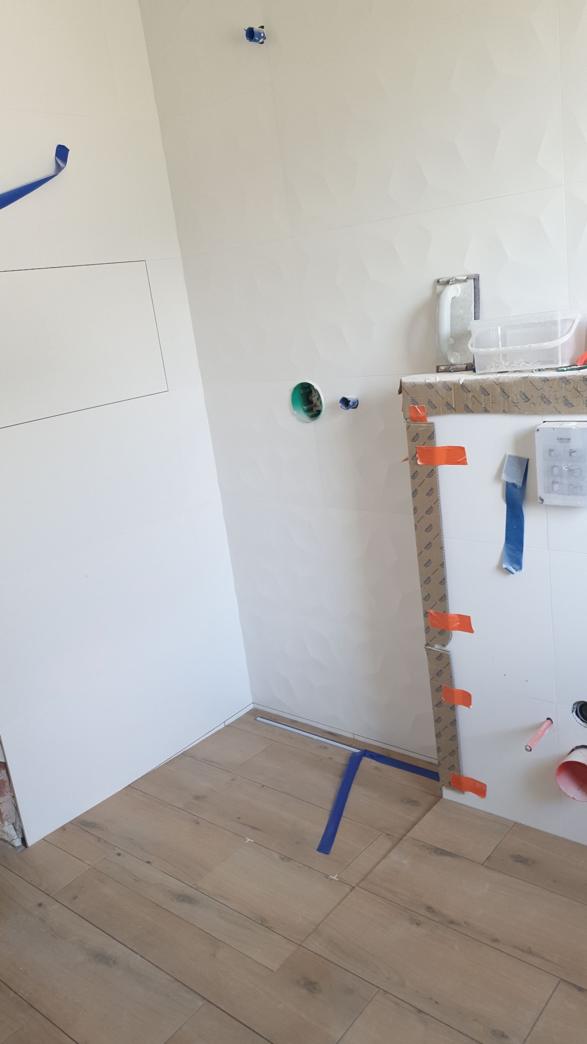 Ułożona podłoga z płytek imitujących drewno w trakcie prac, ściany wyłożone białymi płytkami z geometrycznym wzorem, widoczne otwory na instalacje i niebieska taśma malarska.