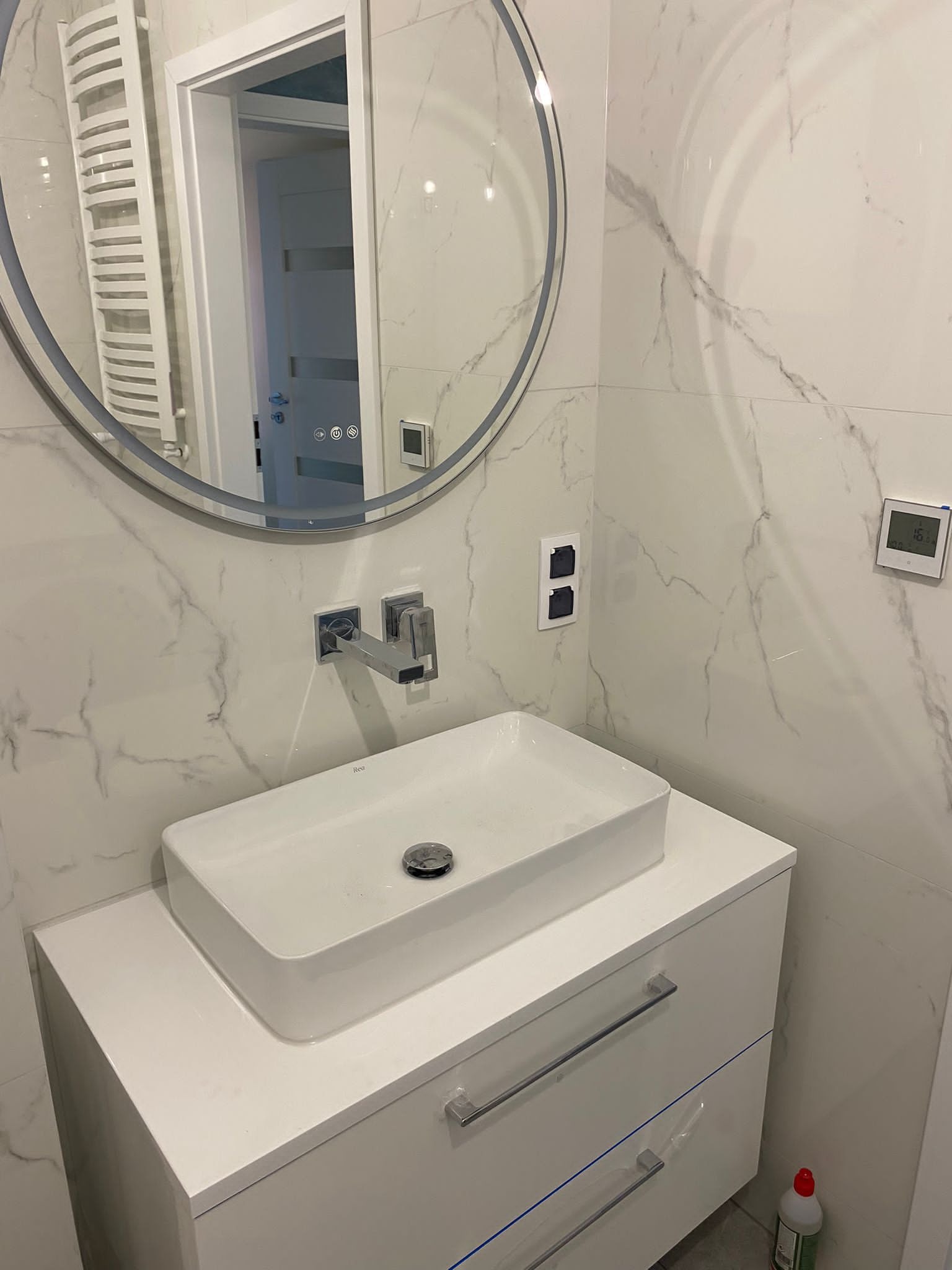 Nowoczesna łazienka z umywalką nablatową, lustrem z podświetleniem LED i marmurowymi płytkami, widoczny termostat i gniazdka elektryczne.