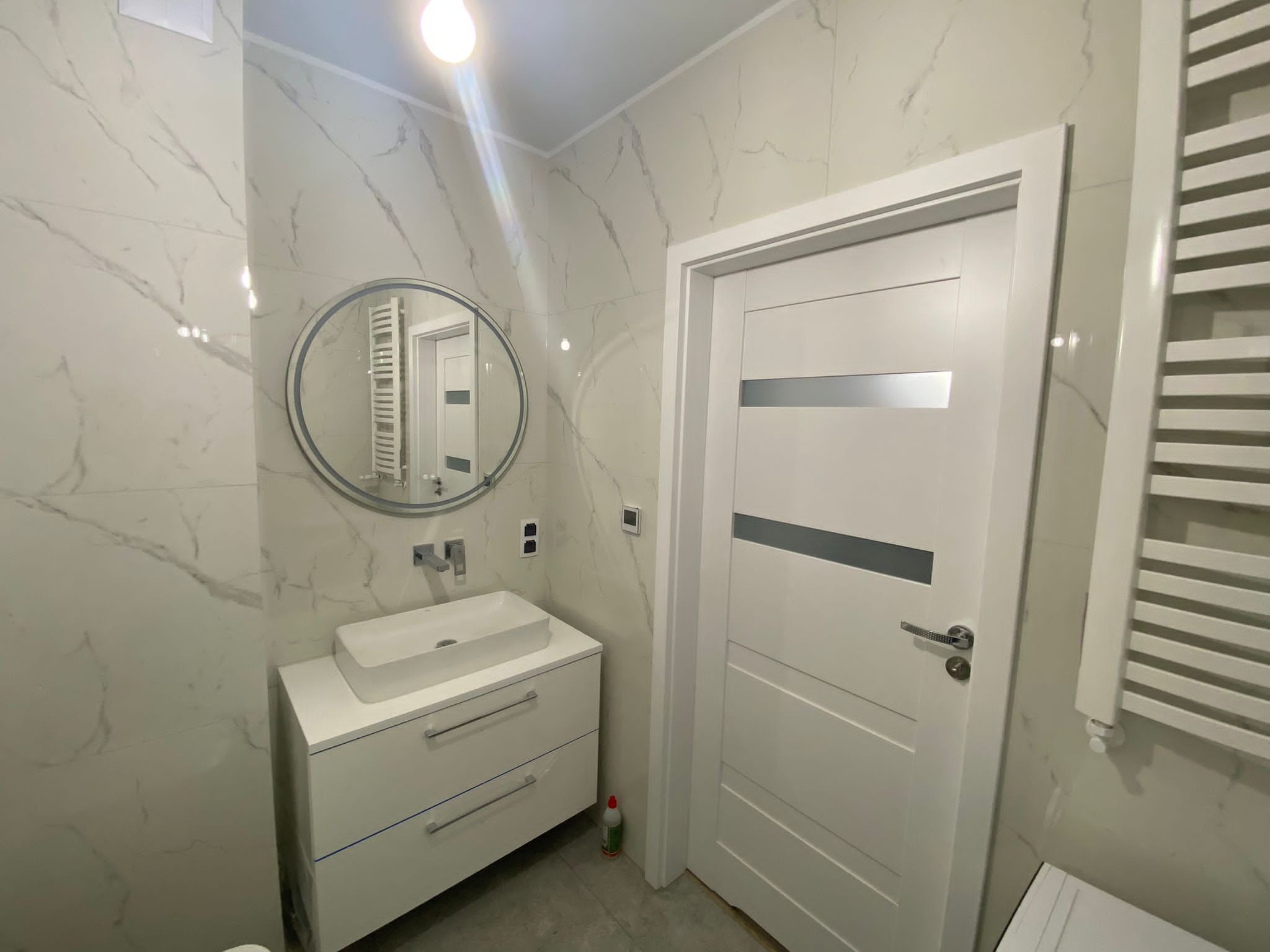 Nowoczesna łazienka z marmurowymi płytkami, owalnym lustrem, białą szafką z umywalką i grzejnikiem drabinkowym, widoczna biała, wewnętrzna ościeżnica drzwiowa z poziomymi szarymi wstawkami.