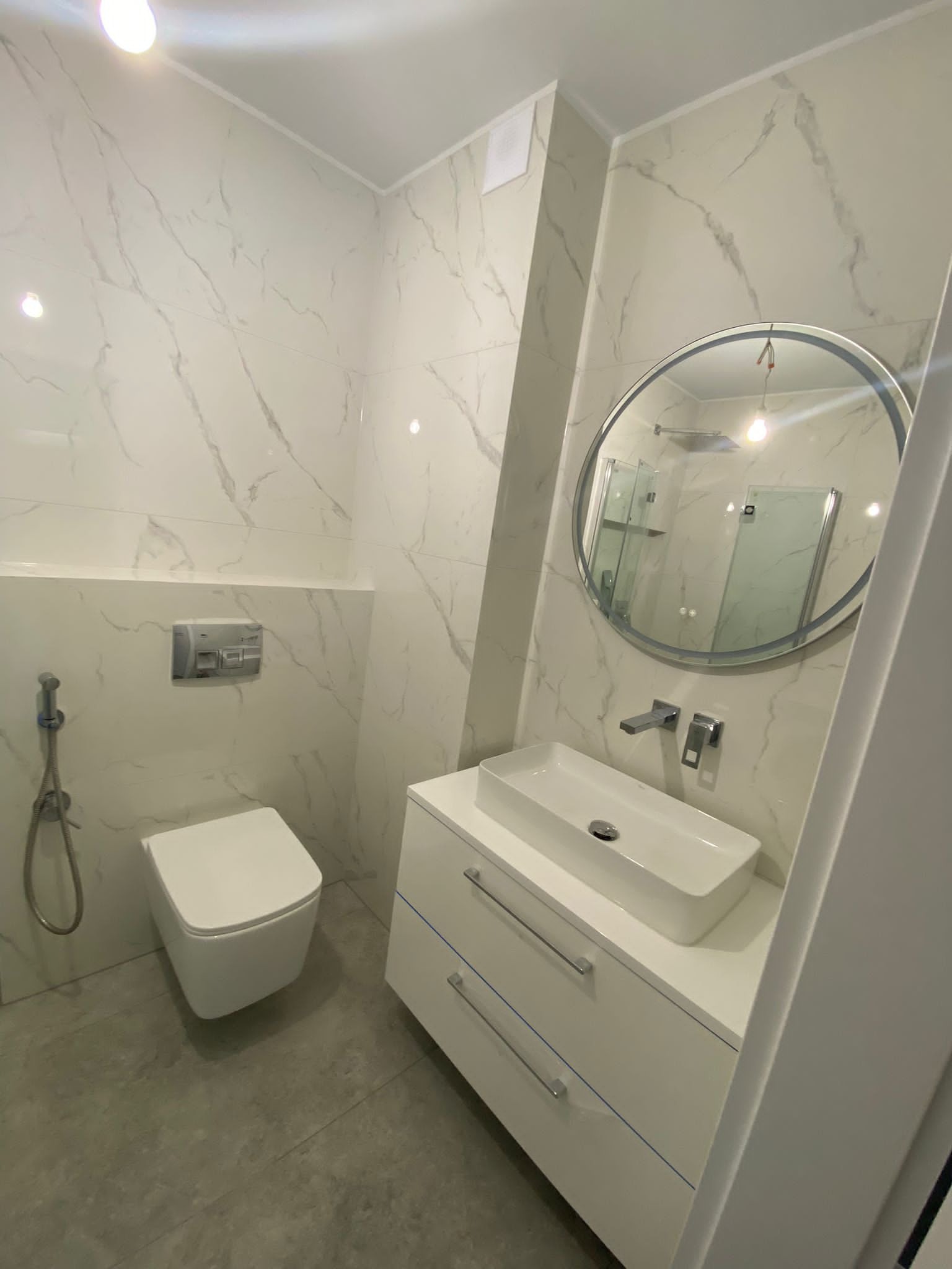 Nowoczesna łazienka z marmurowymi płytkami, podwieszaną toaletą z bidetką, białą szafką z umywalką nablatową i okrągłym lustrem z metalową ramą.