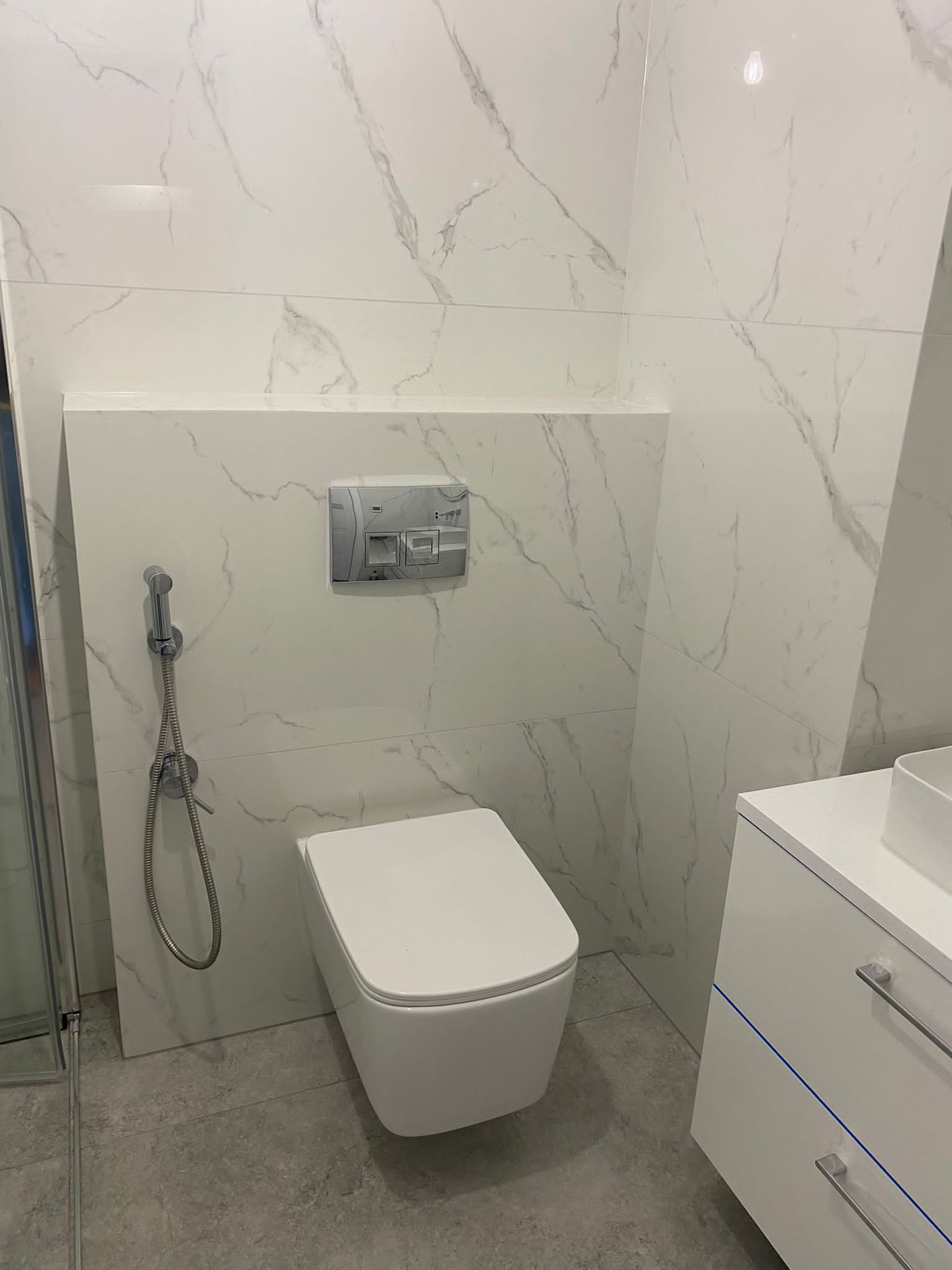 Nowoczesna toaleta podwieszana z bidetką, płytki imitujące marmur na ścianach, szara podłoga i biała szafka umywalkowa z niebieskim akcentem.