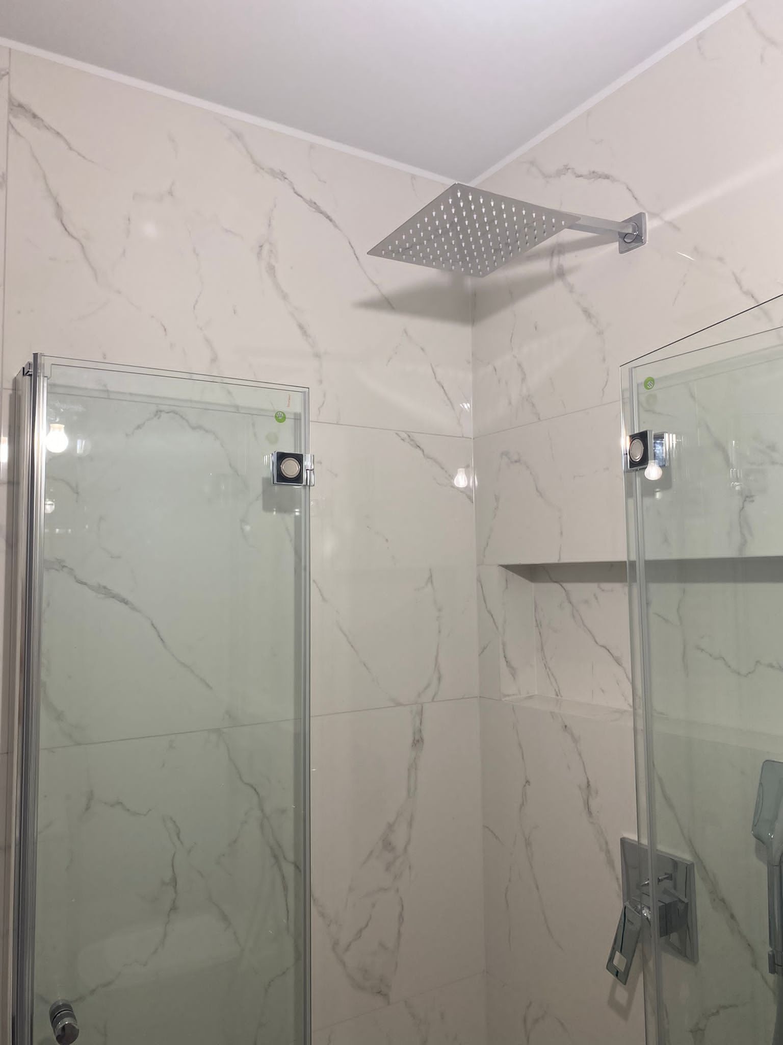 Nowoczesna łazienka z kabiną prysznicową, białe płytki marmurowe, kwadratowa deszczownica chromowana, minimalistyczna armatura, półka wnękowa.