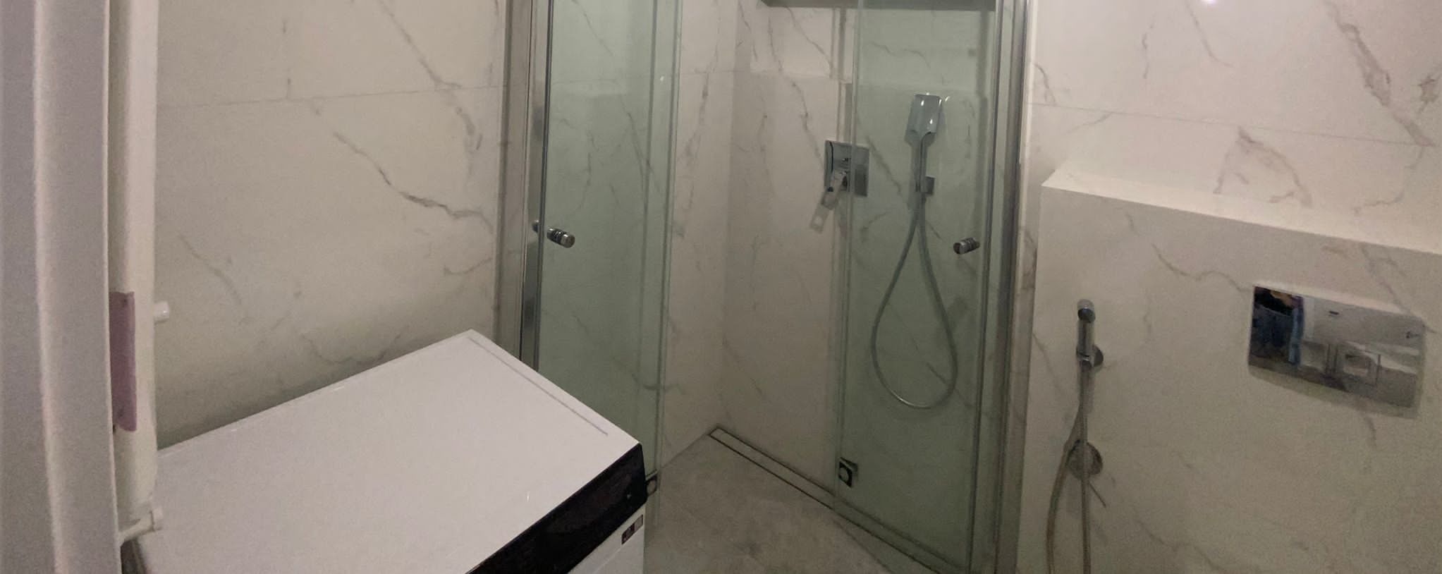 Nowoczesna łazienka z kabiną prysznicową ze szklanymi drzwiami, białe marmurowe płytki na ścianach, biała pralka w rogu, chromowana armatura i odpływ liniowy.