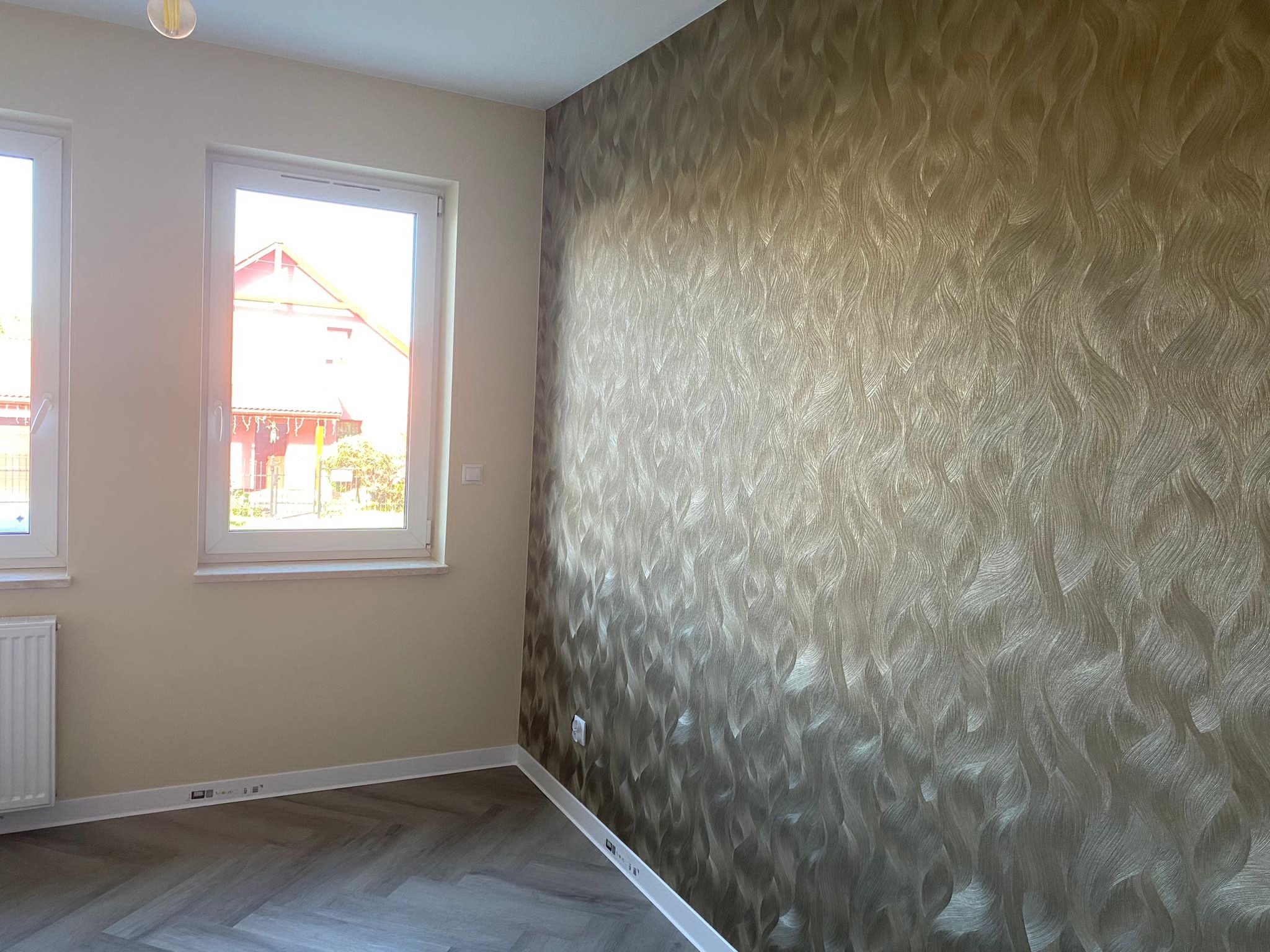 Pokój po remoncie z wzorzystą tapetą w odcieniach beżu i brązu na jednej ścianie, panele podłogowe ułożone w jodełkę, białe okna z widokiem na dom za oknem, biała listwa przypodłogowa.