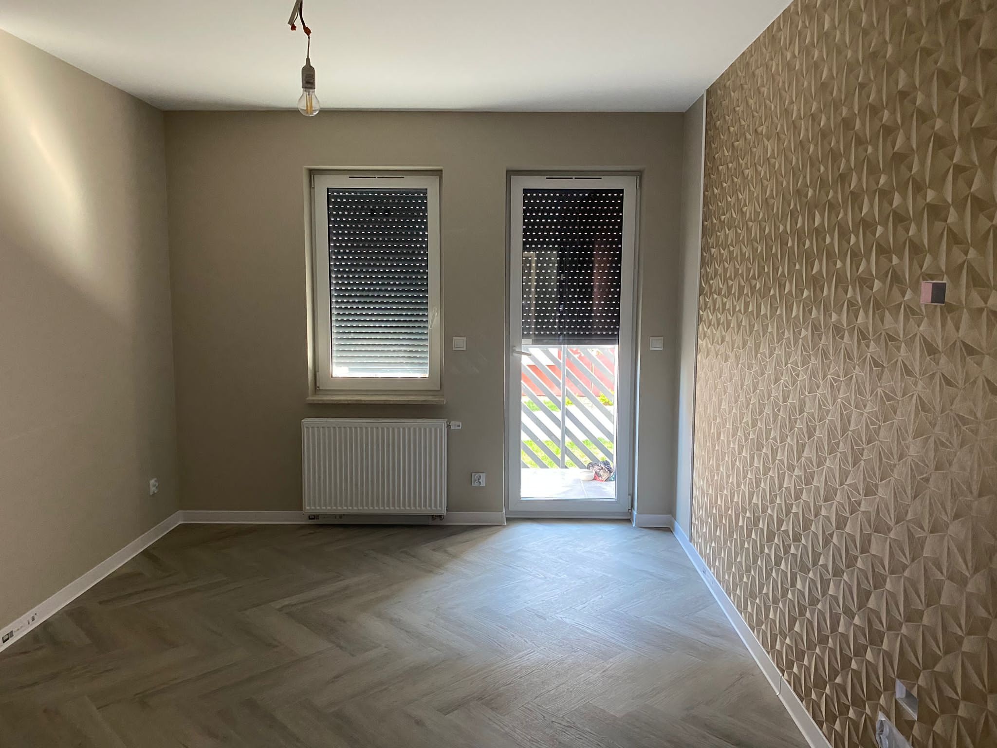 Puste pomieszczenie po remoncie z parkietem w jodełkę, ścianą z trójwymiarową tapetą, grzejnikiem pod oknem z roletami i wiszącą żarówką bez klosza.