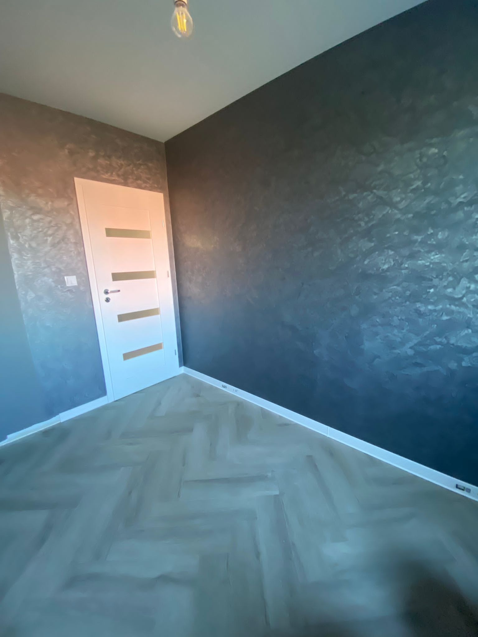 Wnętrze pokoju po remoncie z parkietem ułożonym w jodełkę, ściany pomalowane farbą dekoracyjną z efektem metalicznym, białe drzwi wewnętrzne z poziomymi przeszkleniami, surowa żarówka na suficie.