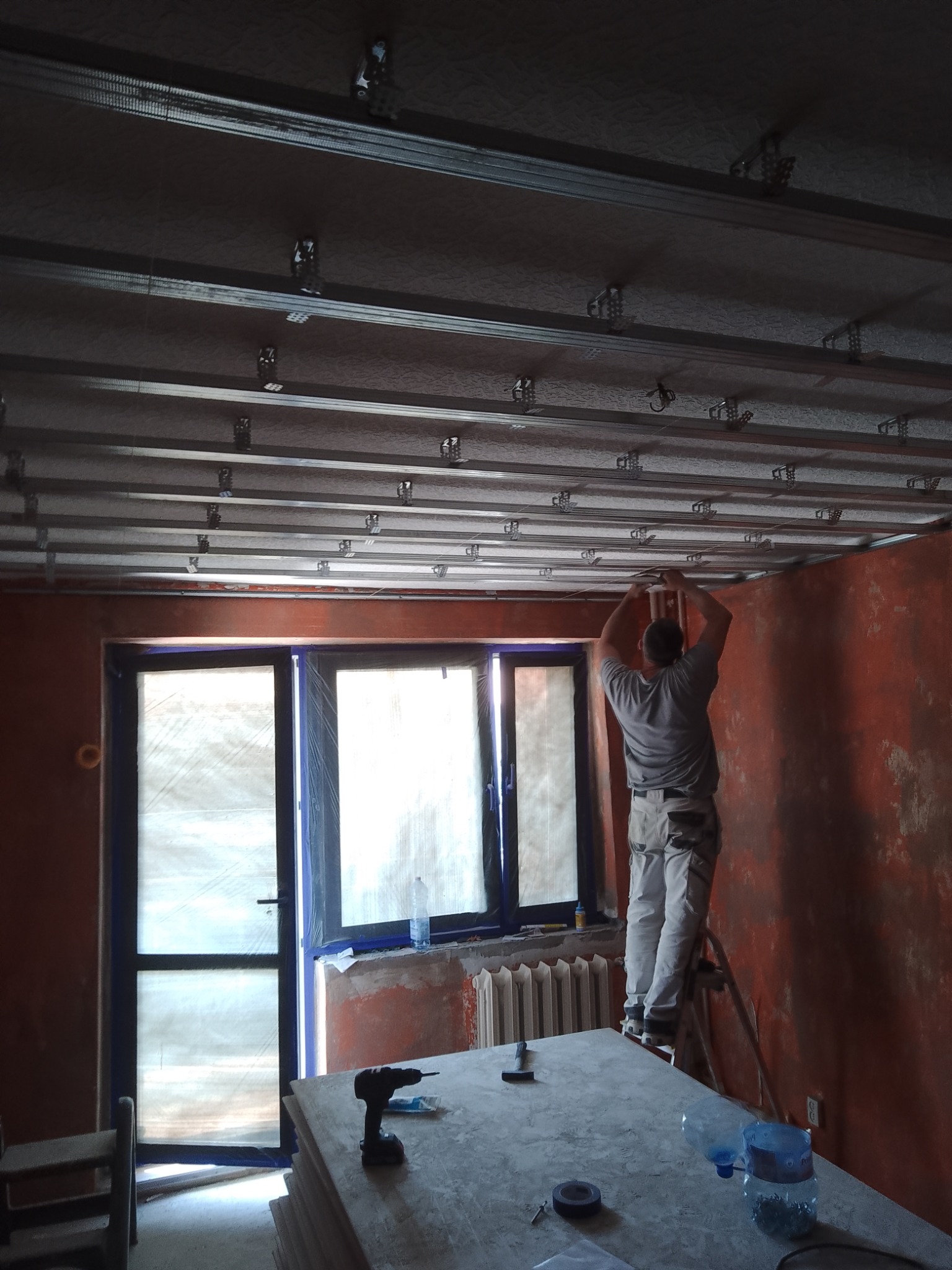 Pracownik na drabinie montuje metalową konstrukcję sufitu podwieszanego z płyt kartonowo-gipsowych w pomieszczeniu z oknem zabezpieczonym folią malarską, widoczny stół roboczy z narzędziami...