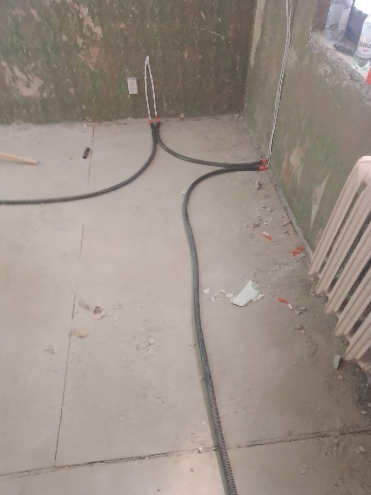 Przygotowanie do instalacji elektrycznej: przewody w peszlach ułożone na surowej podłodze w pomieszczeniu z częściowo odrapanymi ścianami, widoczny fragment grzejnika.