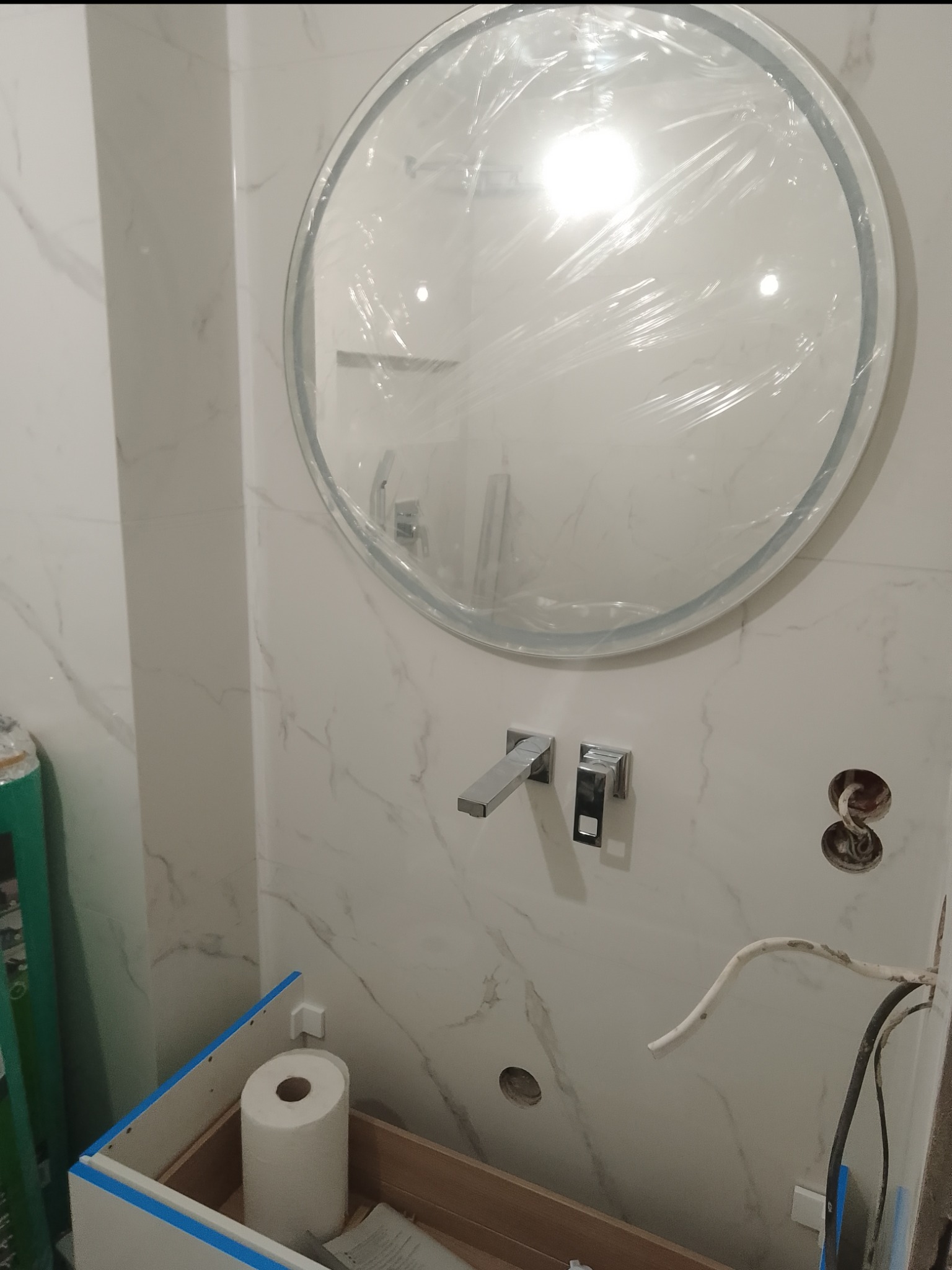 Montaż umywalki w łazience z okrągłym lustrem zabezpieczonym folią, widoczne chromowane bateria i syfon, białe płytki imitujące marmur, oraz instalacja elektryczna w trakcie podłączania.
