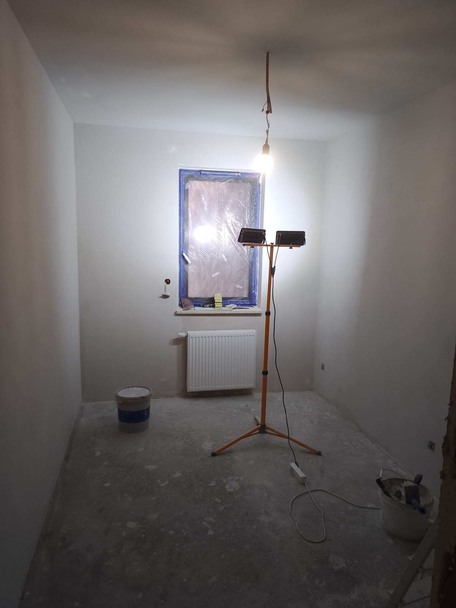 Pusty pokój w trakcie remontu, ściany świeżo pomalowane na biało, okno zabezpieczone folią malarską, na podłodze widoczne ślady farby, stojący reflektor halogenowy oświetla wnętrze, w rogu wiadro...