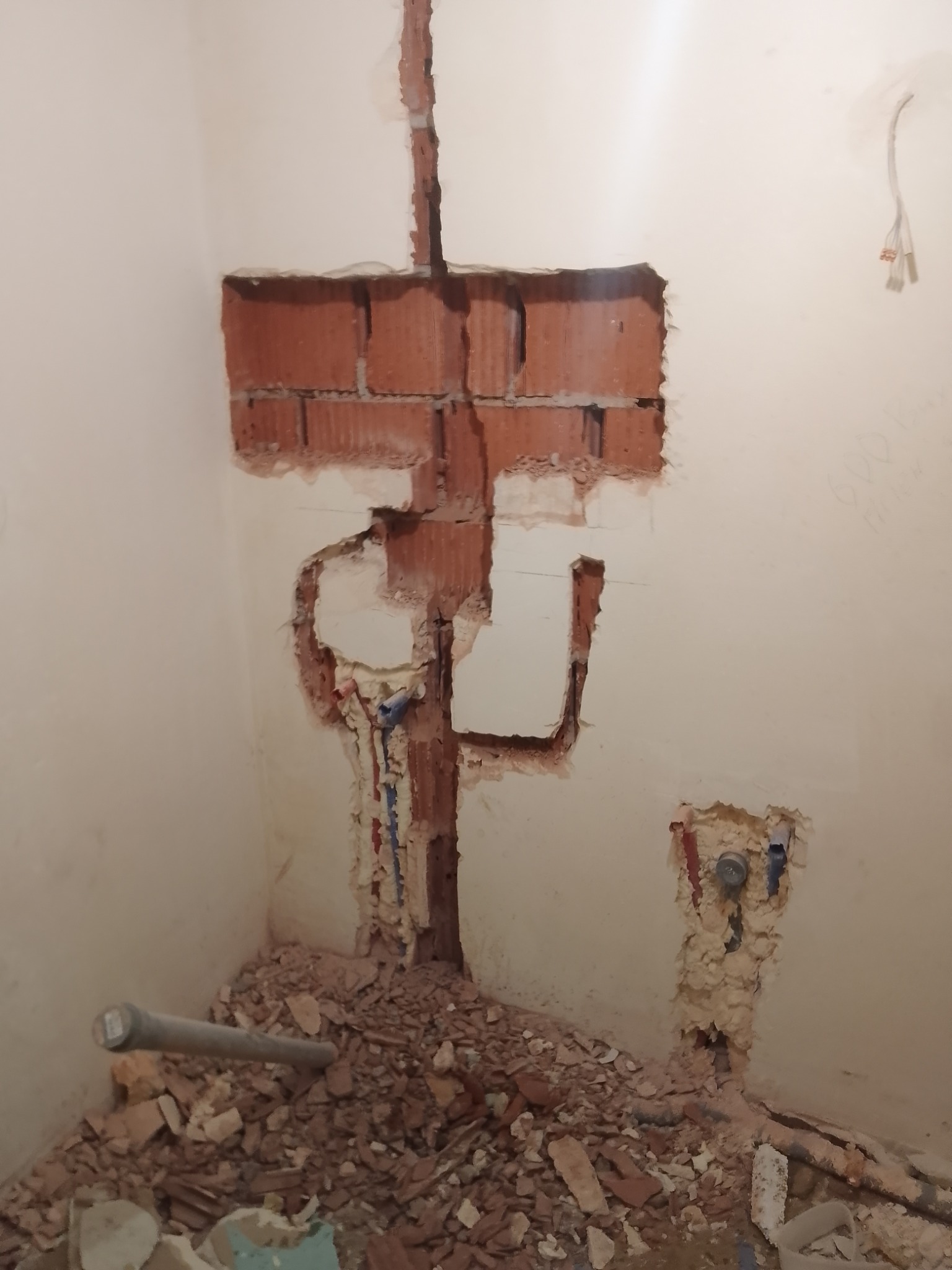 Ściana w trakcie prac instalacyjnych w łazience, z wykuciami pod rury i przewody elektryczne, widoczne fragmenty cegieł i gruz na podłodze.