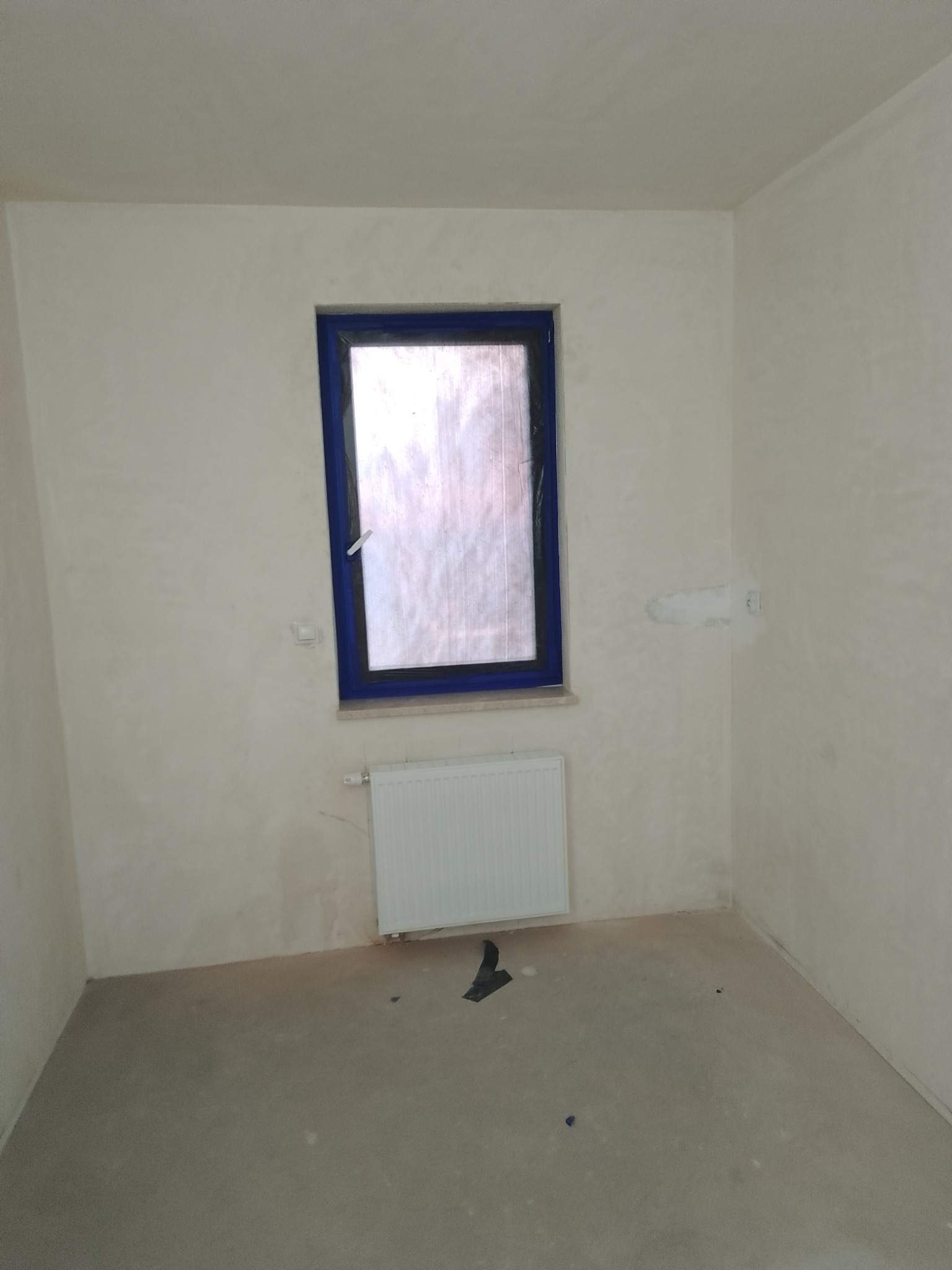 Pokój po nałożeniu gładzi, z niebieską ramą okienną zabezpieczoną folią, biały grzejnik panelowy, surowa podłoga, widoczne ślady po gładzi na ścianach.