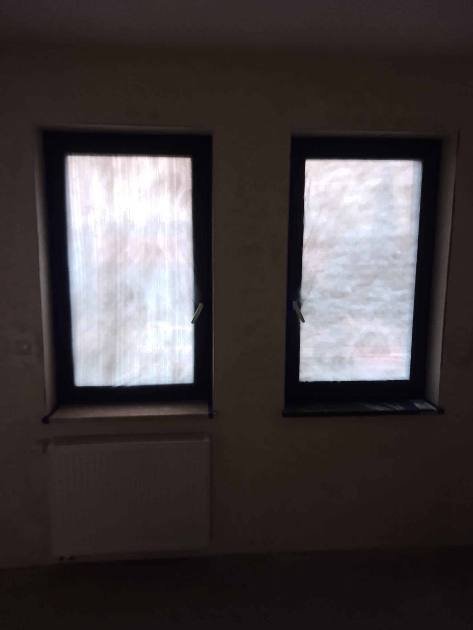Dwa okna z czarnymi ramami zabezpieczone folią malarską, grzejnik pod oknem, ściany przygotowane do malowania, widoczne nierówności i brak wykończenia.
