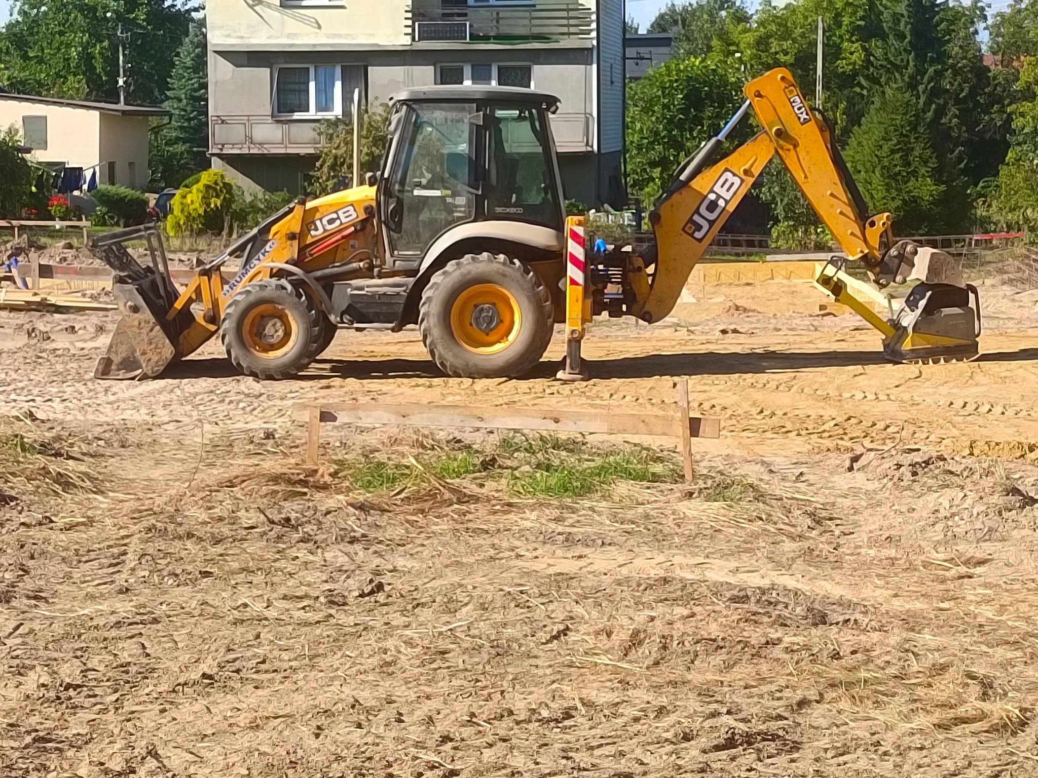 Żółta koparko-ładowarka JCB na placu budowy, przygotowany teren pod fundamenty, drewniane szalunki, w tle budynki mieszkalne i zieleń.