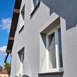 Jacek Hoeft - Szara elewacja domu z brązowymi elementami wykończeniowymi i białymi oknami. Widoczny kolektor słoneczny. Błękitne niebo w tle.