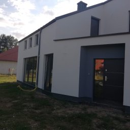 Jacek Hoeft - Nowoczesny dom jednorodzinny z jasną elewacją, ciemnymi oknami i drzwiami. Widoczny fragment ogrodu i sąsiedni budynek z czerwoną dachówką. Ujęcie z boku.