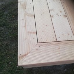 Jacek Hoeft - Fragment drewnianego tarasu z ryflowanych desek, połączonych śrubami, widoczny na tle trawy. Układ desek tworzy kąt prosty, podkreślony listwą wykończeniową.