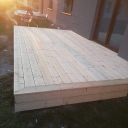 Jacek Hoeft - Nowy, jasny taras drewniany z desek, przylegający do domu z dużym oknem. Widoczne detale łączeń desek i konstrukcji. Słoneczne oświetlenie.