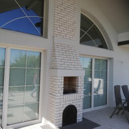 Jacek Hoeft - Biały murowany kominek zewnętrzny na tarasie z szarą kostką, widoczne okna z refleksami nieba i narzędzia budowlane w rogu. Nowoczesny design.