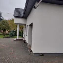 Świeżo otynkowana elewacja domu jednorodzinnego w kolorze złamanej bieli, widoczne okna, filar wejścia, oraz fragment kostki brukowej z rozłożonym przedłużaczem elektrycznym.