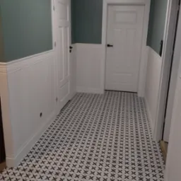 Korytarz z panelami ściennymi w dolnej części i pomalowanymi na zielono ścianami w górnej części, białe drzwi z czarnymi klamkami i wzorzysta podłoga w czarno-białe kwiaty.