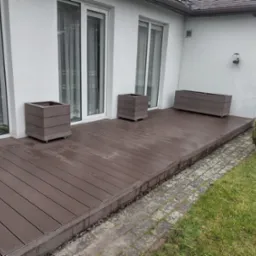 Nowoczesny taras z kompozytu w kolorze ciemnego drewna, z trzema donicami na kółkach, przylegający do białego budynku z dużymi oknami.