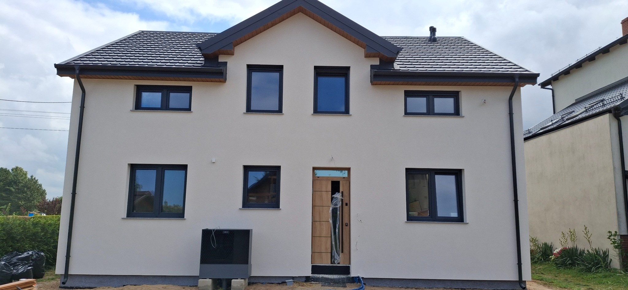 Dwukondygnacyjny dom z jasną elewacją i ciemnymi oknami. Widoczna pompa ciepła przy ścianie. Nowoczesny styl, świeżo po remoncie.