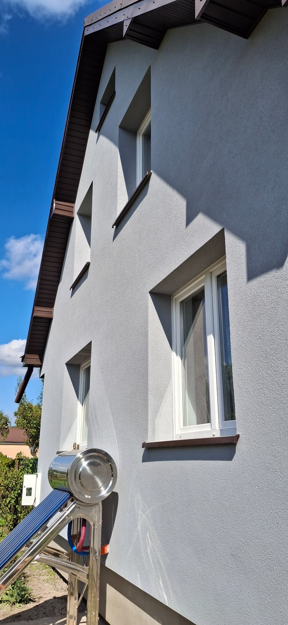 Szara elewacja domu z brązowymi elementami wykończeniowymi i białymi oknami. Widoczny kolektor słoneczny. Błękitne niebo w tle.