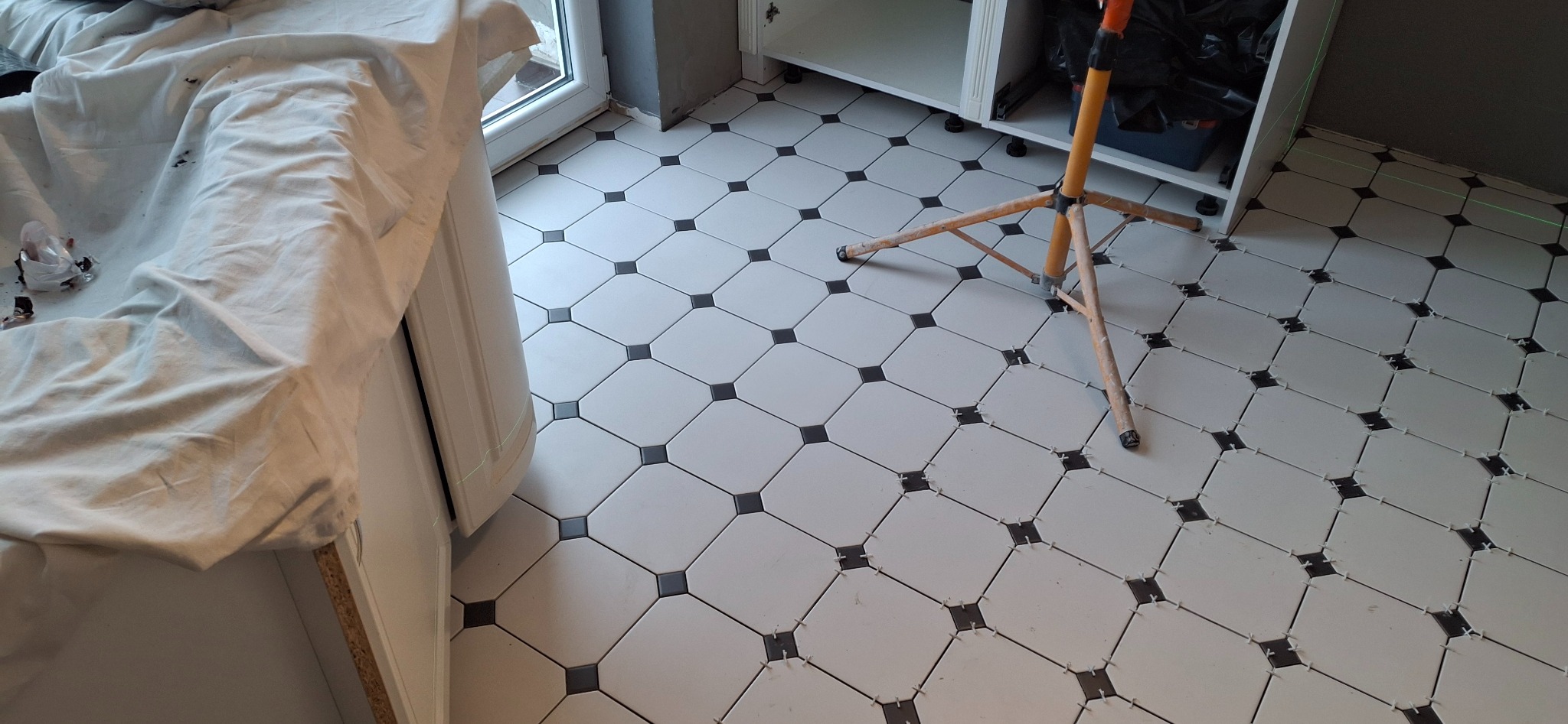 Ułożone białe płytki podłogowe z czarnymi akcentami, widoczne krzyżyki dystansowe, poziomica laserowa i fragment szafki. Układanie płytek w trakcie realizacji.