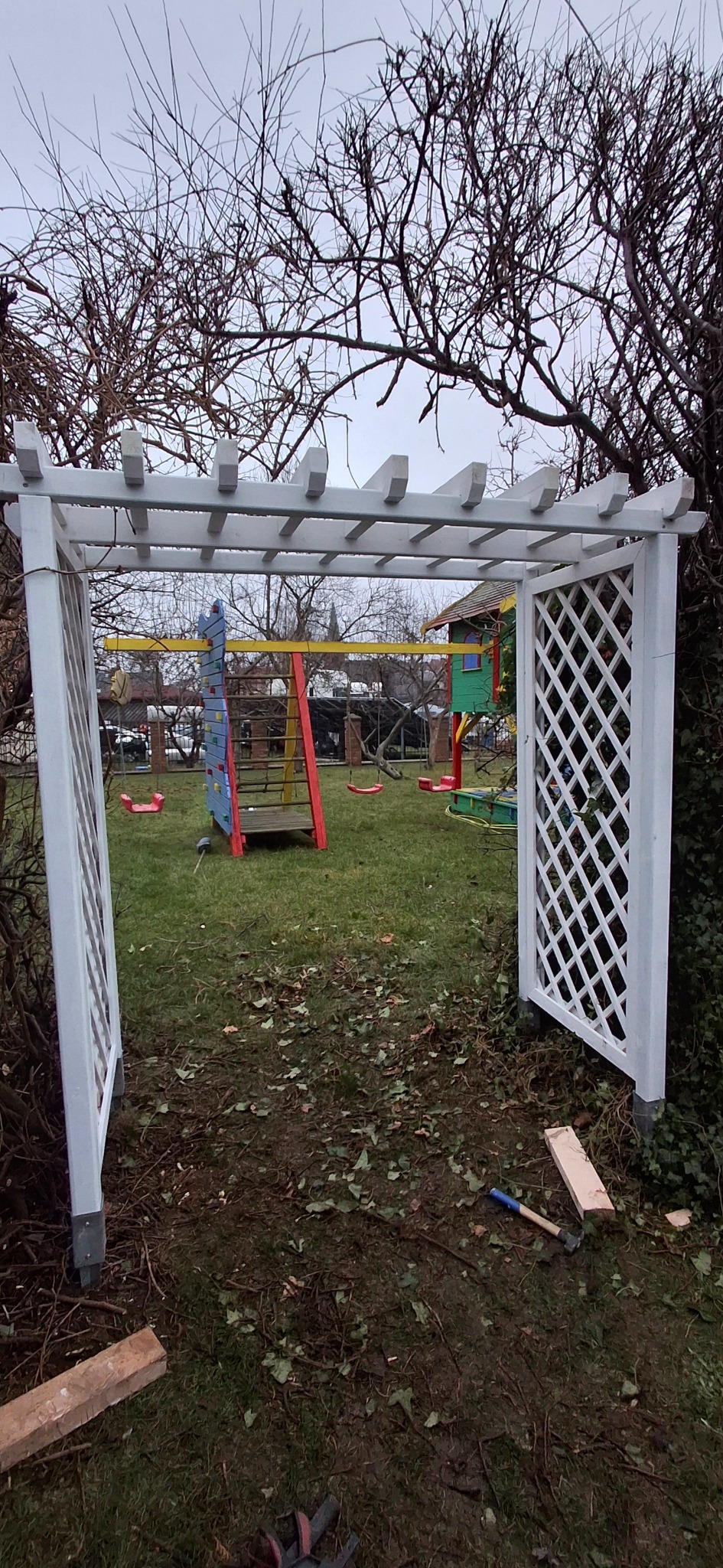 Biała pergola z kratownicą w ogrodzie, widok na plac zabaw. Elementy drewniane i narzędzia sugerują montaż lub prace wykończeniowe.
