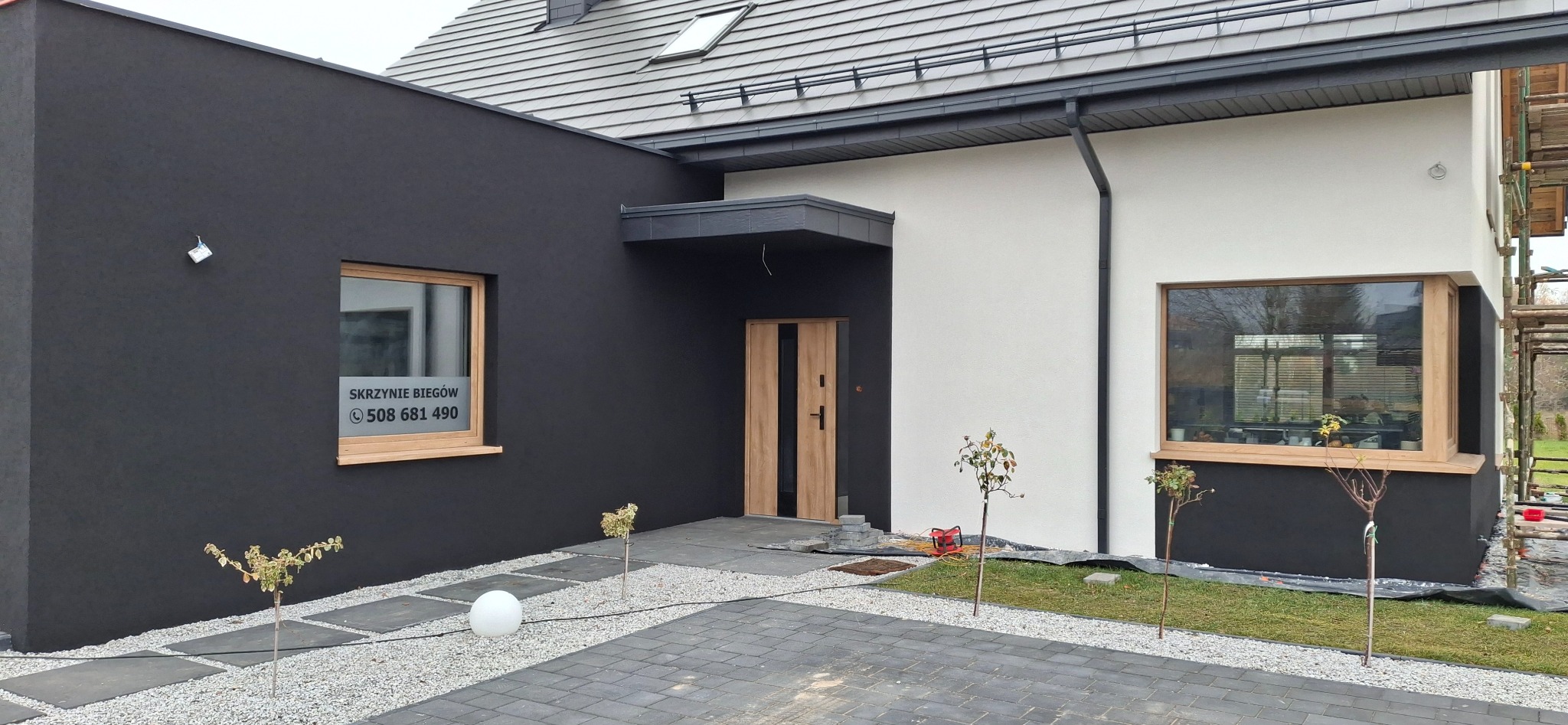 Nowoczesny dom z elewacją w kolorach bieli i grafitu, drewnianą stolarką okienną i drzwiową. Uwagę zwraca minimalistyczny ogródek z kamieniami i młodymi drzewkami.