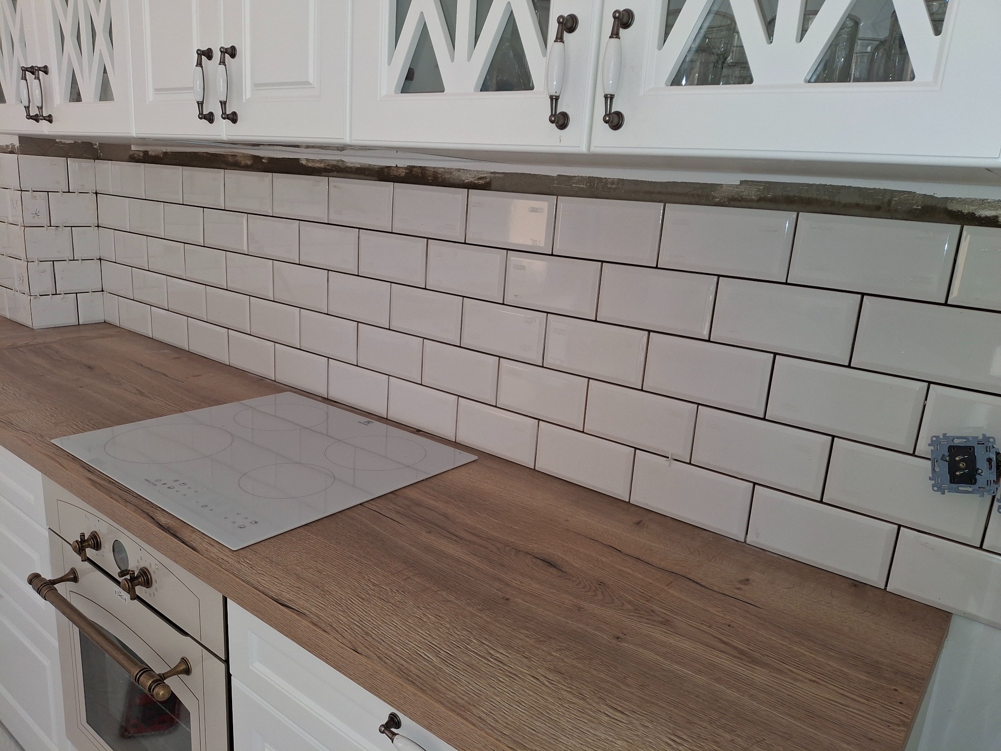 Białe, prostokątne płytki ułożone na ścianie nad blatem imitującym drewno. Widoczna płyta indukcyjna i fragment białej zabudowy kuchennej z ozdobnymi uchwytami.