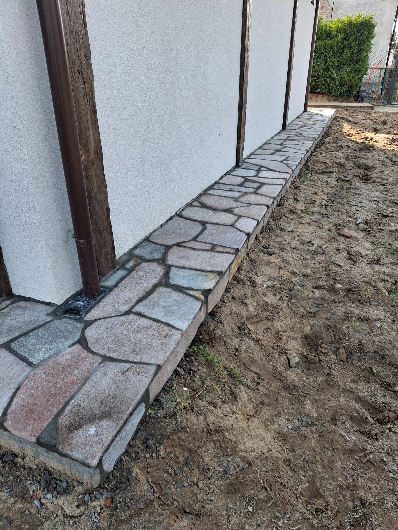 Kamienna opaska wokół domu w Łęczycach, wykonana z nieregularnych płyt. Widoczna rynna i fragment elewacji. Estetyczne wykończenie.