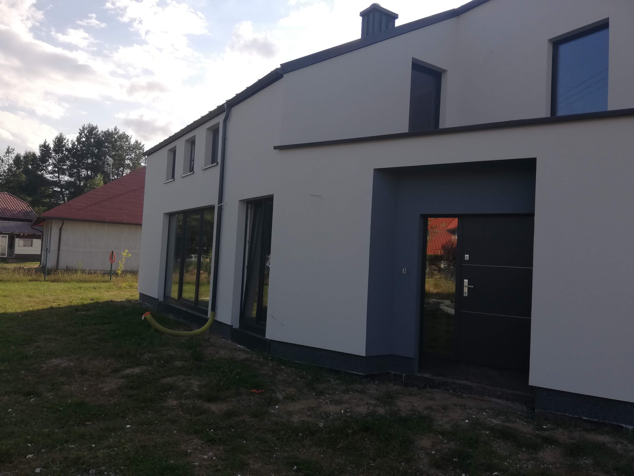 Nowoczesny dom jednorodzinny z jasną elewacją, ciemnymi oknami i drzwiami. Widoczny fragment ogrodu i sąsiedni budynek z czerwoną dachówką. Ujęcie z boku.