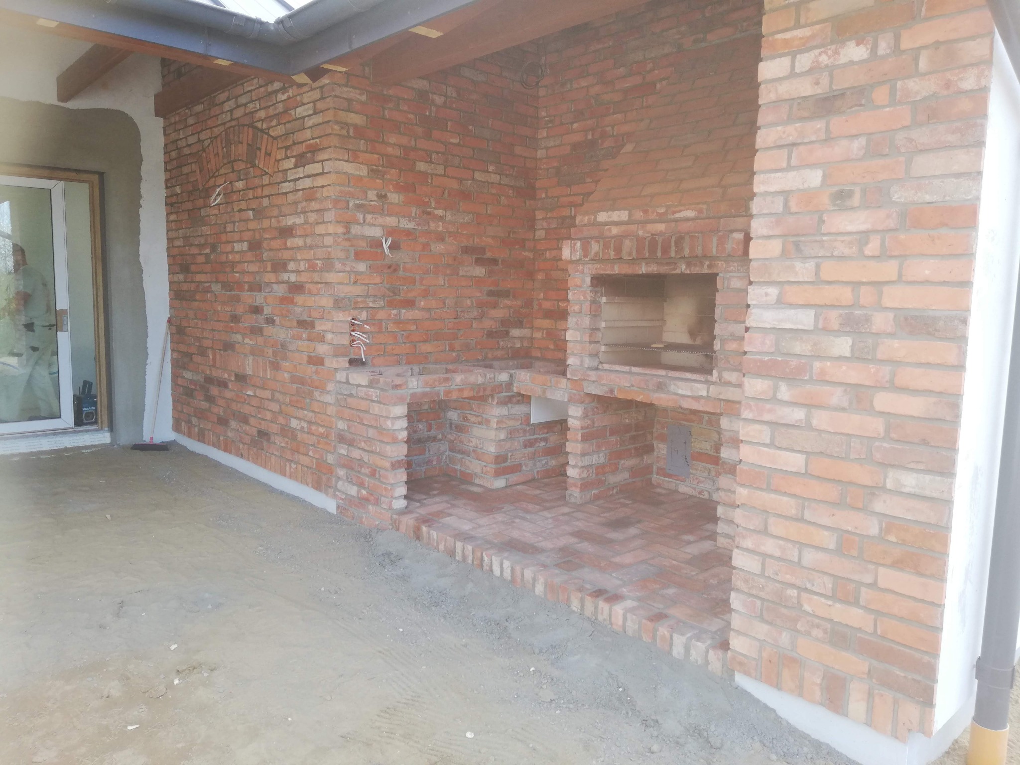 Murowana z cegły klinkierowej zabudowa grilla z wnękami na akcesoria, zadaszona konstrukcja, surowy stan posadzki, widok z boku.