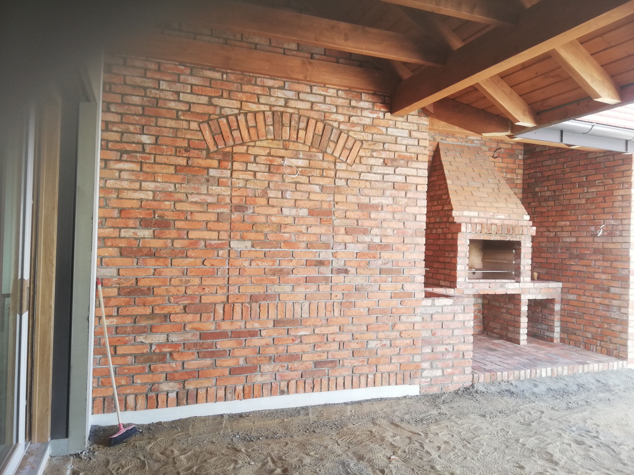 Murowany grill z cegły klinkierowej pod drewnianą konstrukcją zadaszenia. Widoczna ściana z cegły z łukowym zdobieniem. Na pierwszym planie zamiatarka.