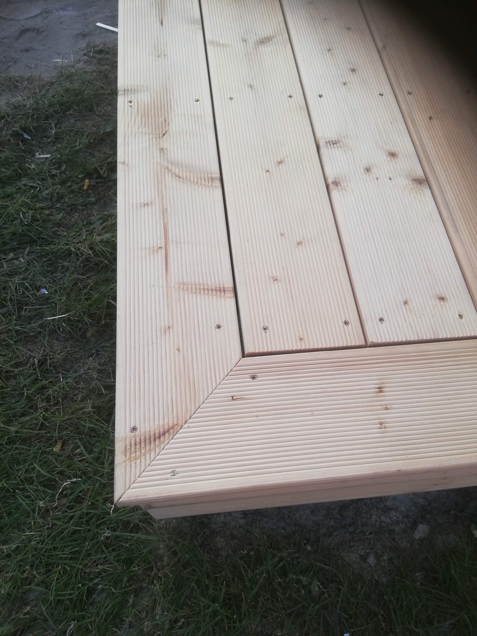 Fragment drewnianego tarasu z ryflowanych desek, połączonych śrubami, widoczny na tle trawy. Układ desek tworzy kąt prosty, podkreślony listwą wykończeniową.