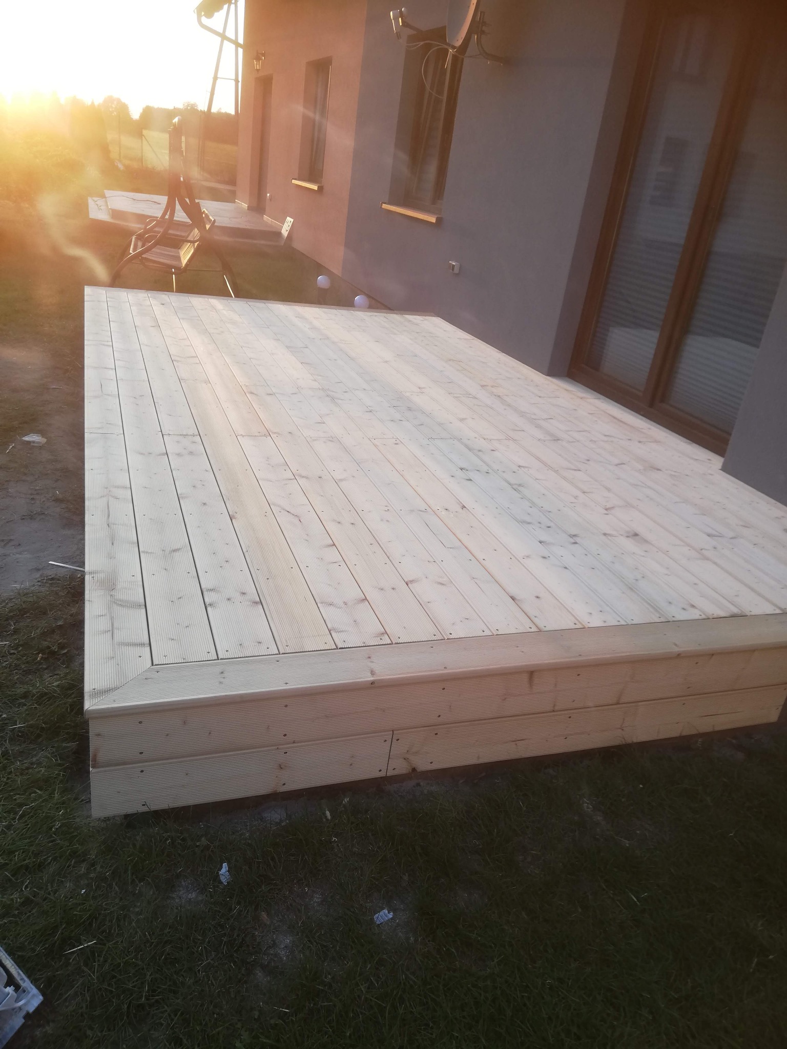 Nowy, jasny taras drewniany z desek, przylegający do domu z dużym oknem. Widoczne detale łączeń desek i konstrukcji. Słoneczne oświetlenie.