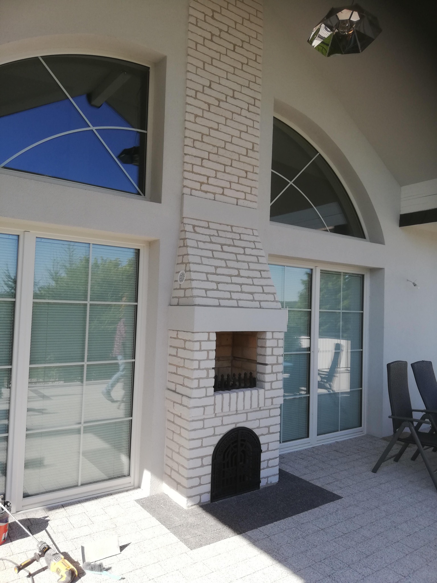 Biały murowany kominek zewnętrzny na tarasie z szarą kostką, widoczne okna z refleksami nieba i narzędzia budowlane w rogu. Nowoczesny design.