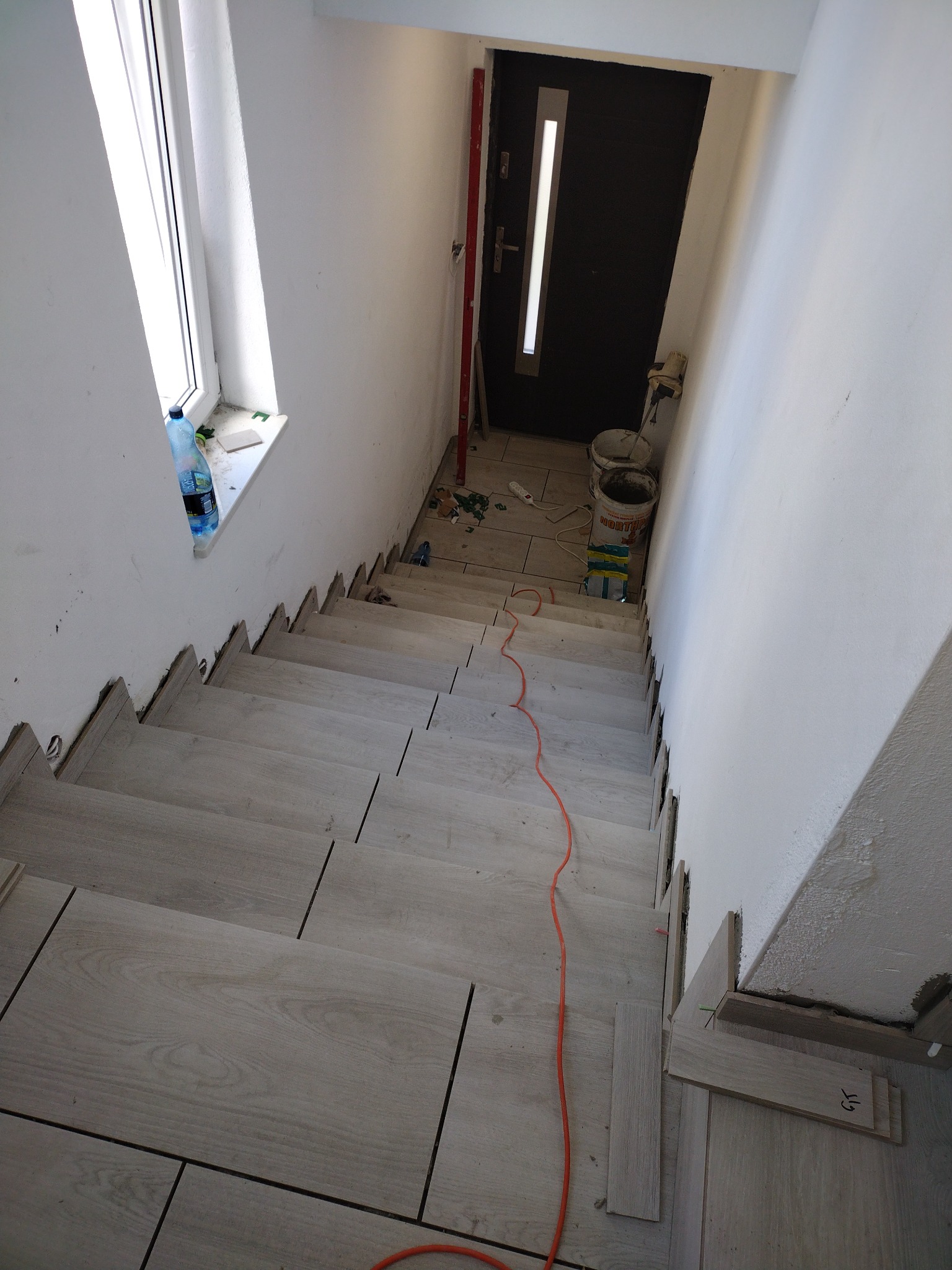 Montaż płytek imitujących drewno na schodach wewnętrznych; widoczne białe ściany, okno z butelką wody, drzwi wejściowe, poziomica i materiały budowlane.