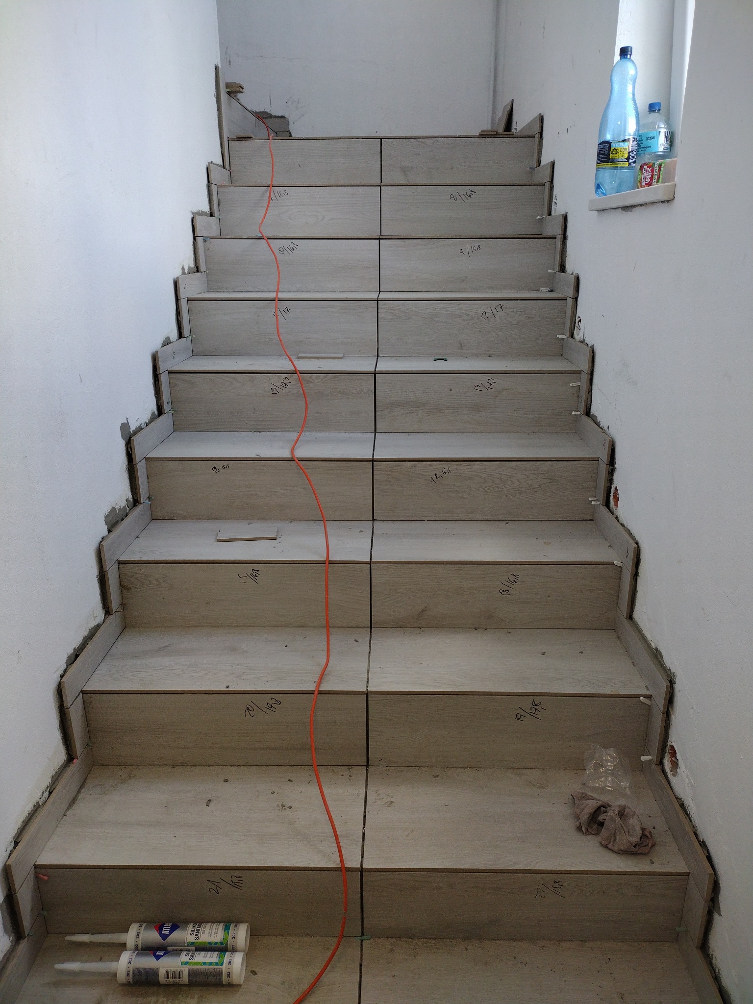 Montaż paneli na schodach w trakcie realizacji, widoczne przycięte panele, silikon, kabel i notatki na panelach.