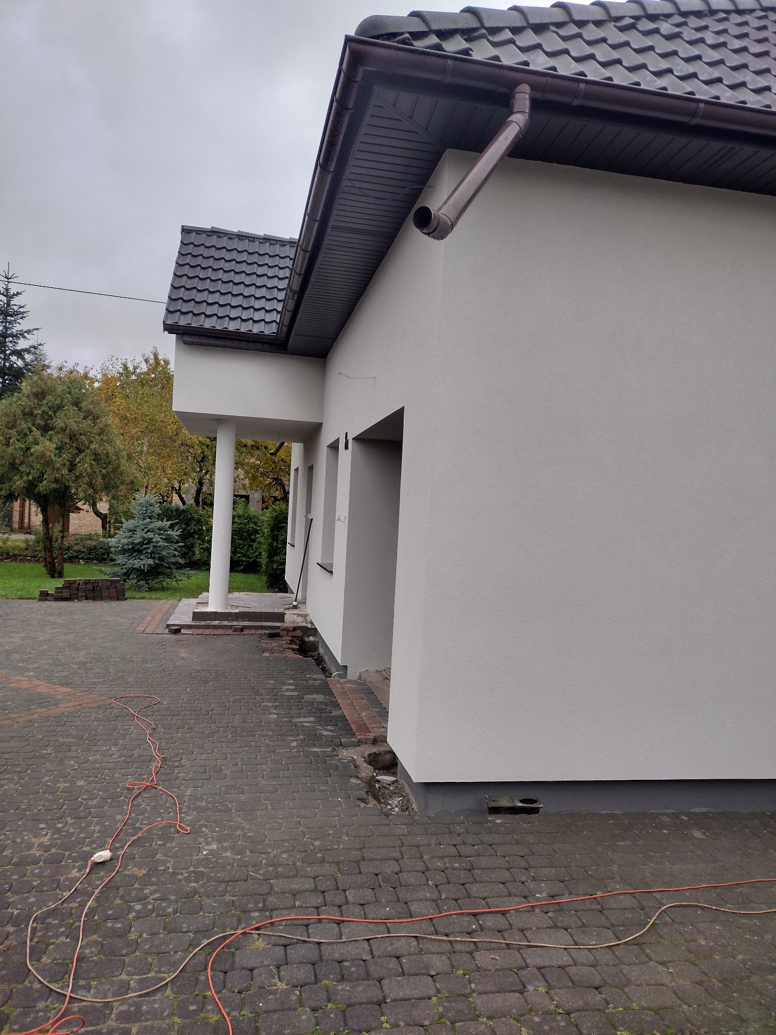 Świeżo otynkowana elewacja domu jednorodzinnego w kolorze złamanej bieli, widoczne okna, filar wejścia, oraz fragment kostki brukowej z rozłożonym przedłużaczem elektrycznym.