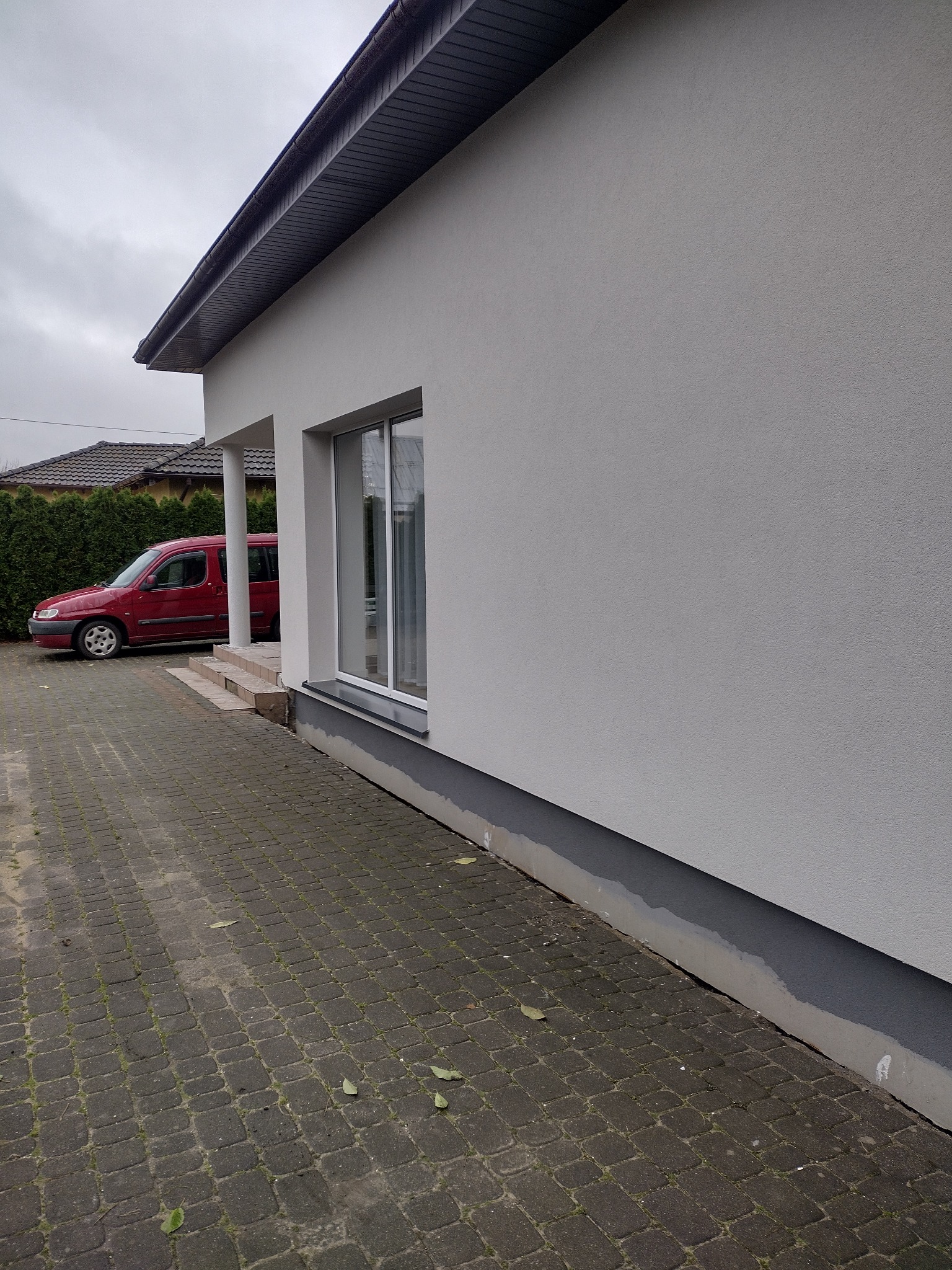 Świeżo otynkowana, jasna elewacja domu jednorodzinnego z widocznym fragmentem okna i szarego cokołu, na tle brukowanego podjazdu i zaparkowanego czerwonego samochodu.