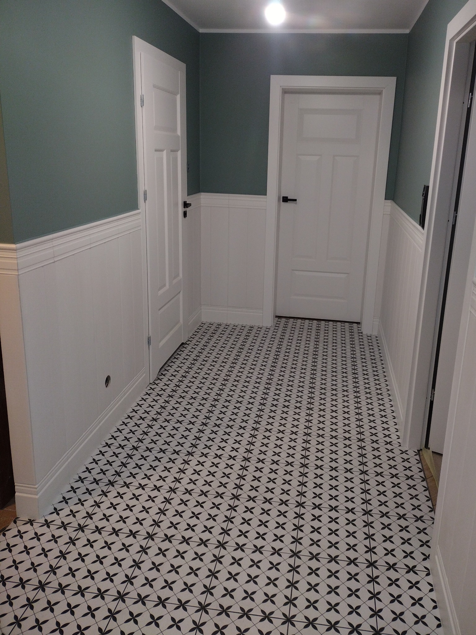 Korytarz z panelami ściennymi w dolnej części i pomalowanymi na zielono ścianami w górnej części, białe drzwi z czarnymi klamkami i wzorzysta podłoga w czarno-białe kwiaty.