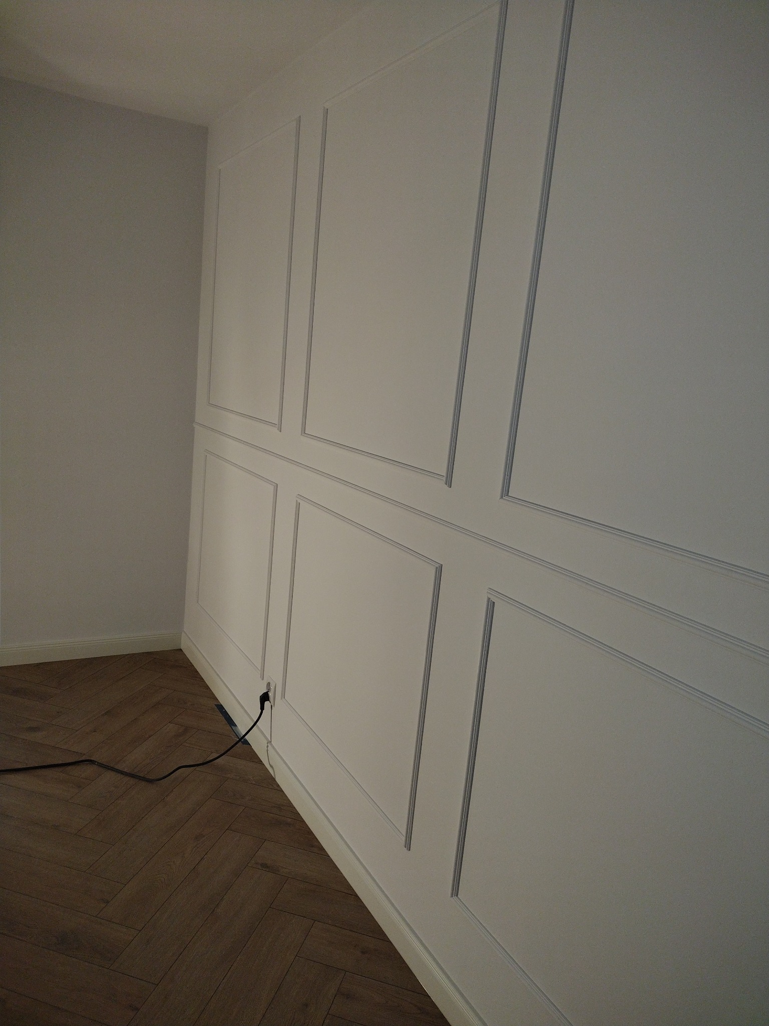Biała ściana z dekoracyjnymi panelami w stylu angielskim, widoczny fragment parkietu ułożonego w jodełkę i kabel zasilający wystający ze ściany.