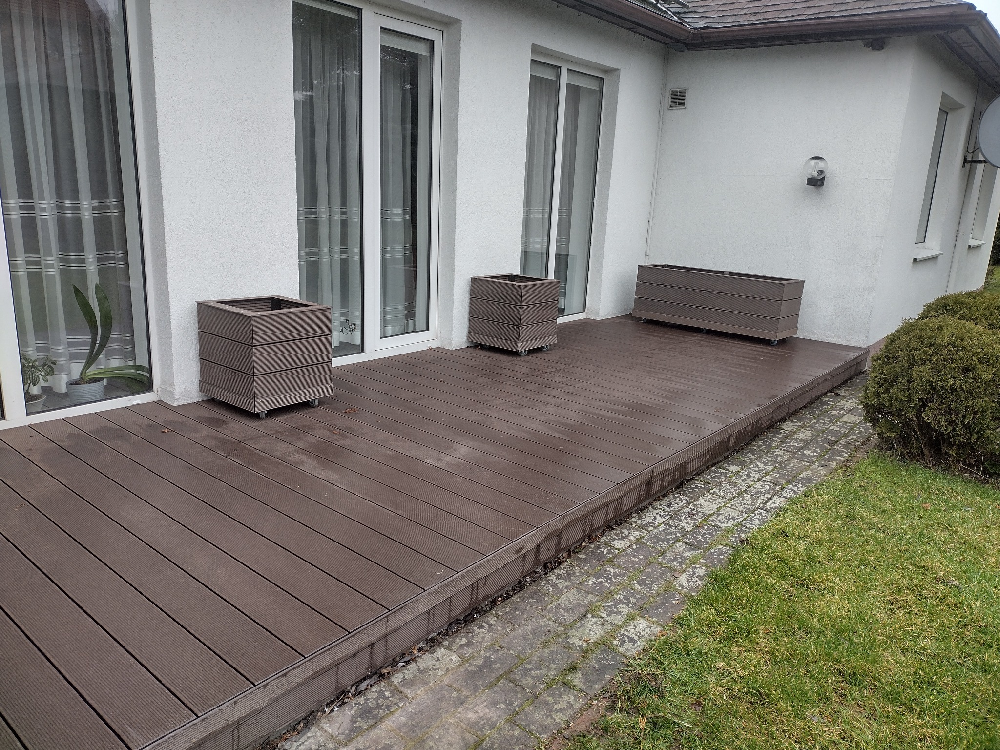 Nowoczesny taras z kompozytu w kolorze ciemnego drewna, z trzema donicami na kółkach, przylegający do białego budynku z dużymi oknami.