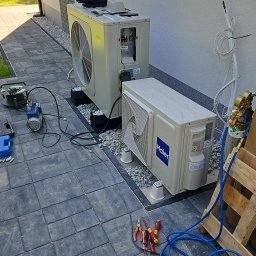 Novatech Energy sp. z o.o. - Dwie jednostki zewnętrzne klimatyzacji, jedna marki Midea, druga Haier, połączone przewodami, na jednej z nich urządzenie pomiarowe Testo, obok rozłożone narzędzia i butla z gazem, na kostce brukowej.